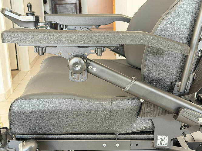 Nahaufnahme einer verstellbaren Armlehne an einem gepolsterten Stuhl mit Metallmechanik. Nicht eindeutig erkennbar aber könnte sich um einen elektrischen Rollstuhl handeln.