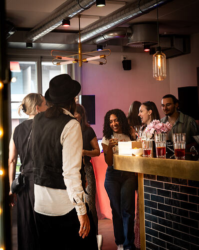 Mehrere Personen stehen an einer Bar, unterhalten sich und trinken, auf der Theke stehen Gläser und Blumen.
