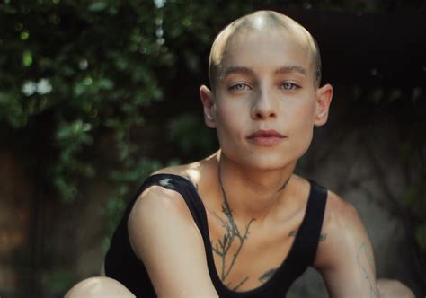 Eine Person mit rasiertem Kopf und hellen Augen sitzt in natürlichem Licht, umgeben von grünem Laub. Feine Tattoos an Hals und Schlüsselbein betonen ihren individuellen Stil. Sie trägt ein schwarzes Top und blickt direkt in die Kamera.