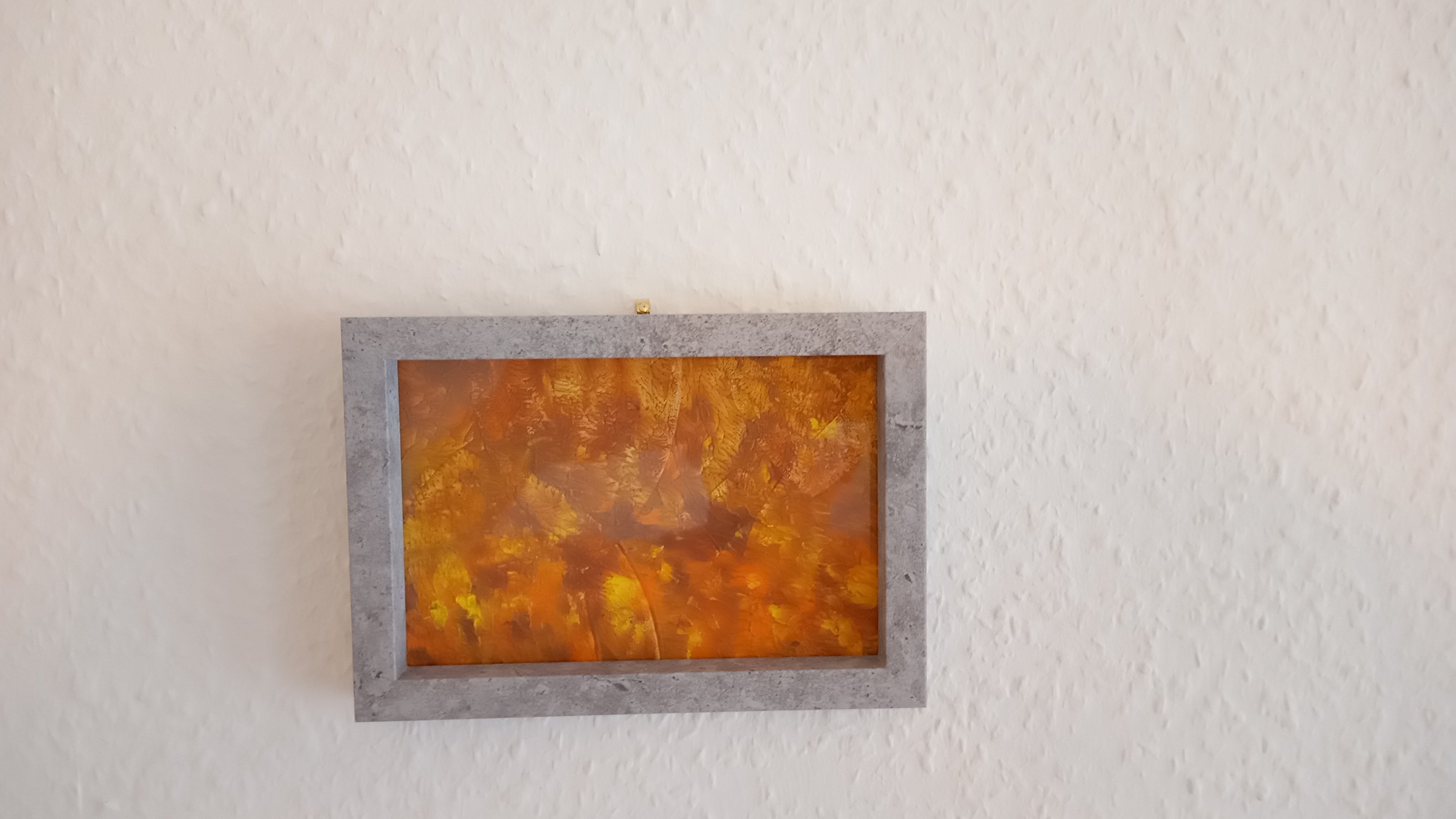 Grauer Bilderrahmen auf weisser Wand. Der Künstler hat verschiedene Wachsmalfarben auf ein Papier geraspelt und mit einem Bügeleisen zum Schmelzen gebracht. Es sind hauptsächlich die Farben Hell-Orange, Dunkel-Orange und Gelb.