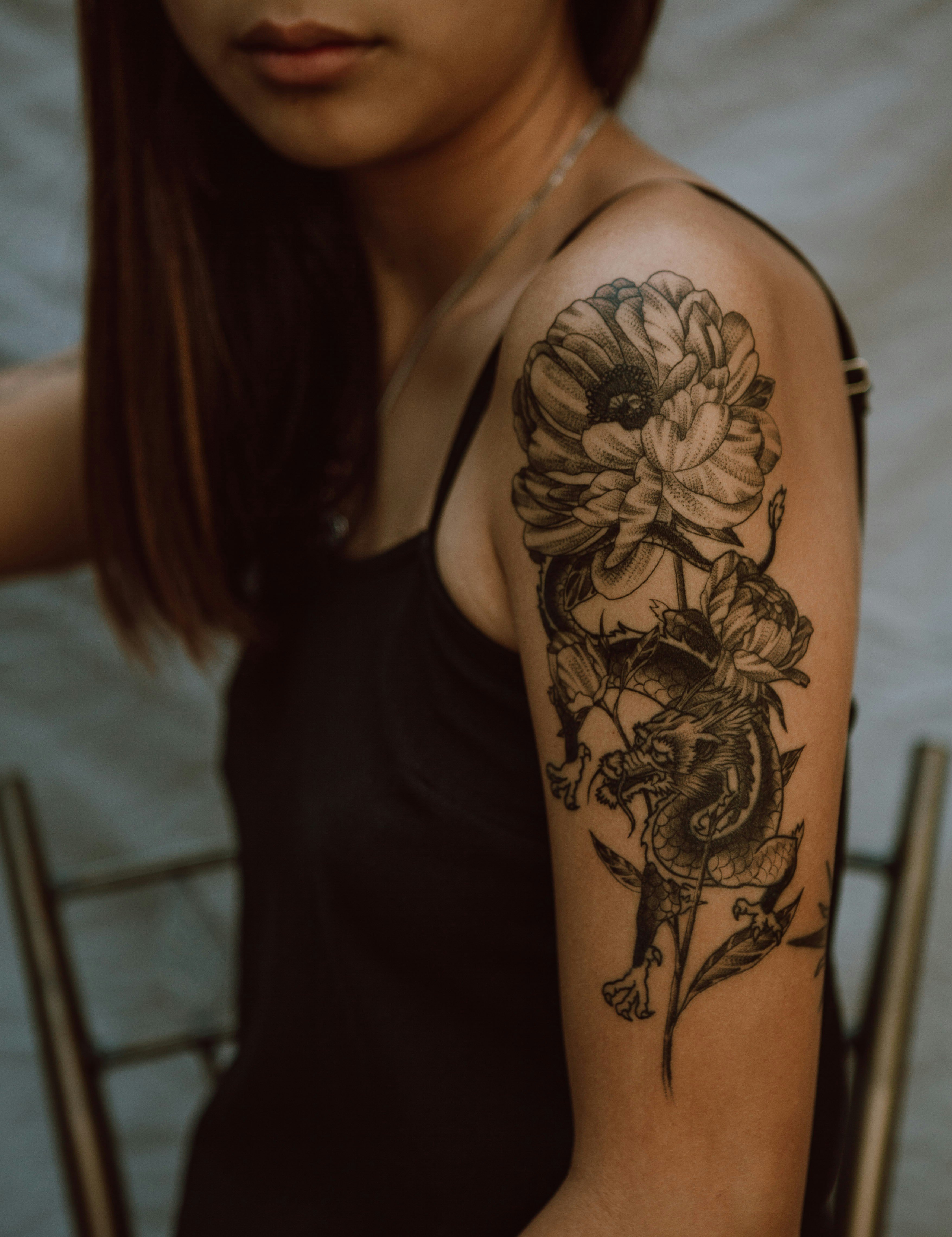 Ein Tattoo auf dem Oberarm einer Frau. Das Tattoo zeigt ein detailliertes Design mit einer großen Blume, die wie eine Chrysantheme aussieht, und einem Drachen, der sich um die Blume windet. Die Linien sind fein und das gesamte Design ist in Schwarz gehalten. Die Frau trägt ein schwarzes ärmelloses Oberteil und hat lange, glatte Haare. Der Hintergrund ist unscharf, sodass der Fokus auf dem Tattoo liegt.