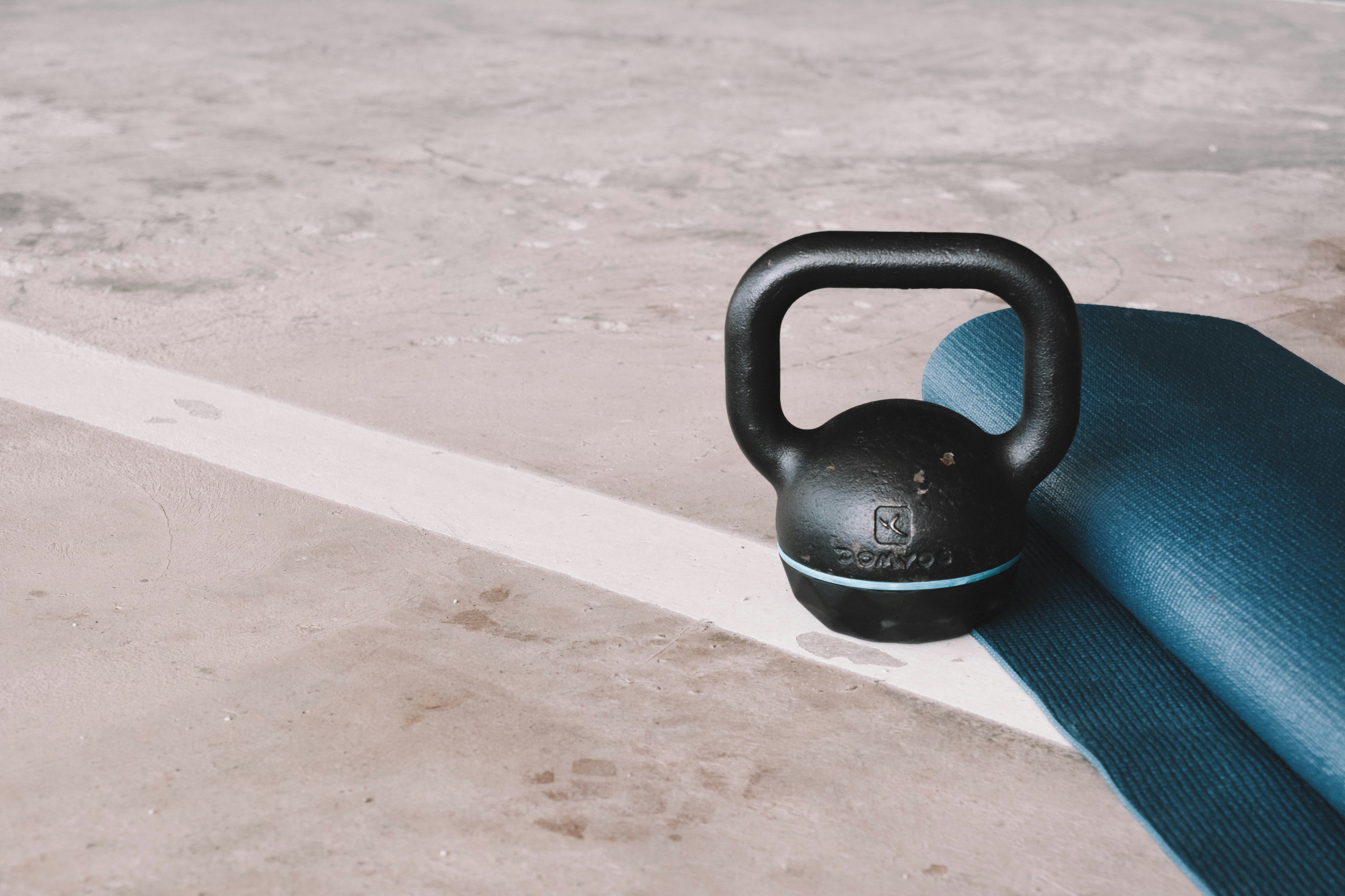 Eine Kettlebell und eine Yogamatte. Auf dem Bild ist eine schwarze Kettlebell mit einem blauen Streifen zu sehen, die auf einem Betonboden steht. Daneben liegt eine zusammengerollte blaue Yogamatte. Der Boden hat eine weiße Linie, die diagonal durch das Bild verläuft. Die Kettlebell hat eine Aufschrift, die schwer zu erkennen ist.