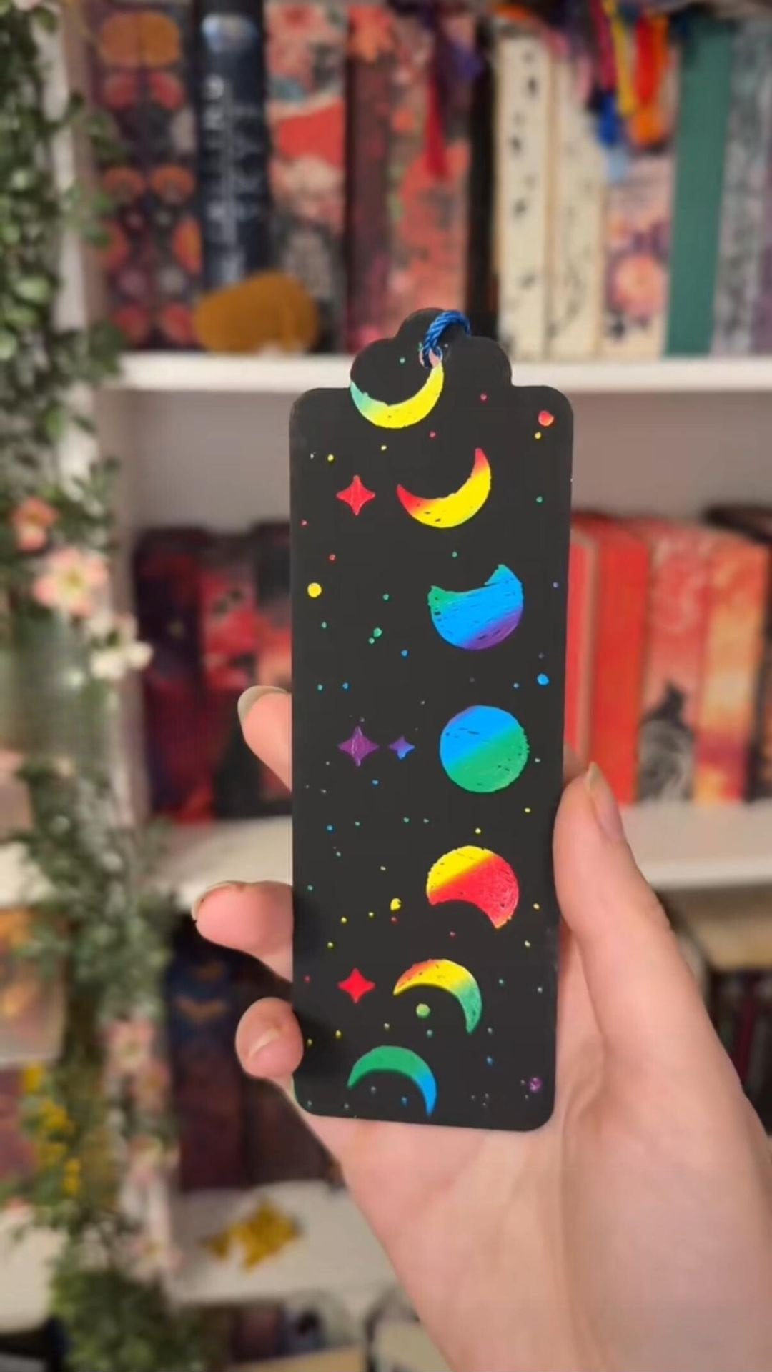Ein schwarzes Lesezeichen hat ein Design aus bunten Mondsicheln und Sternen in Regenbogenfarben. Die Mondsicheln sind in verschiedenen Phasen dargestellt und verlaufen von oben nach unten. Im Hintergrund sind verschwommen Bücherregale zu sehen.