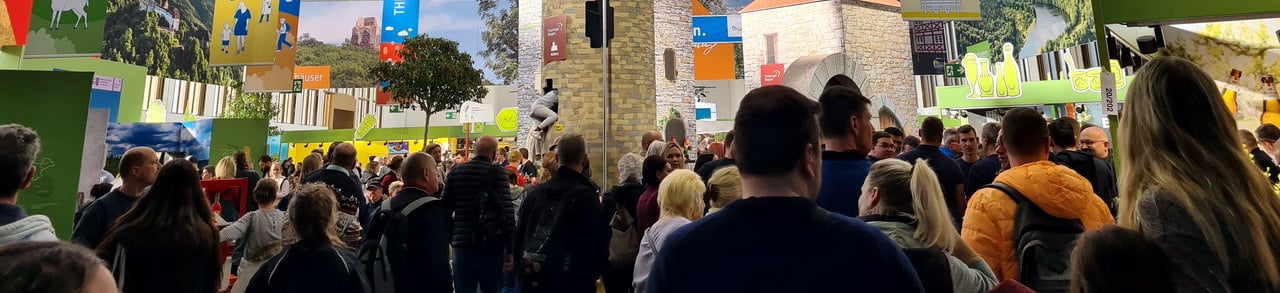Ein Foto einer Messehalle von Innen. Es sind sehr viele Menschen zu sehen und die Halle sieht sehr voll aus. Generell ist es ein sehr buntes Bild, durch die vielen verschiedenen Banner, die aufgehängt wurden.