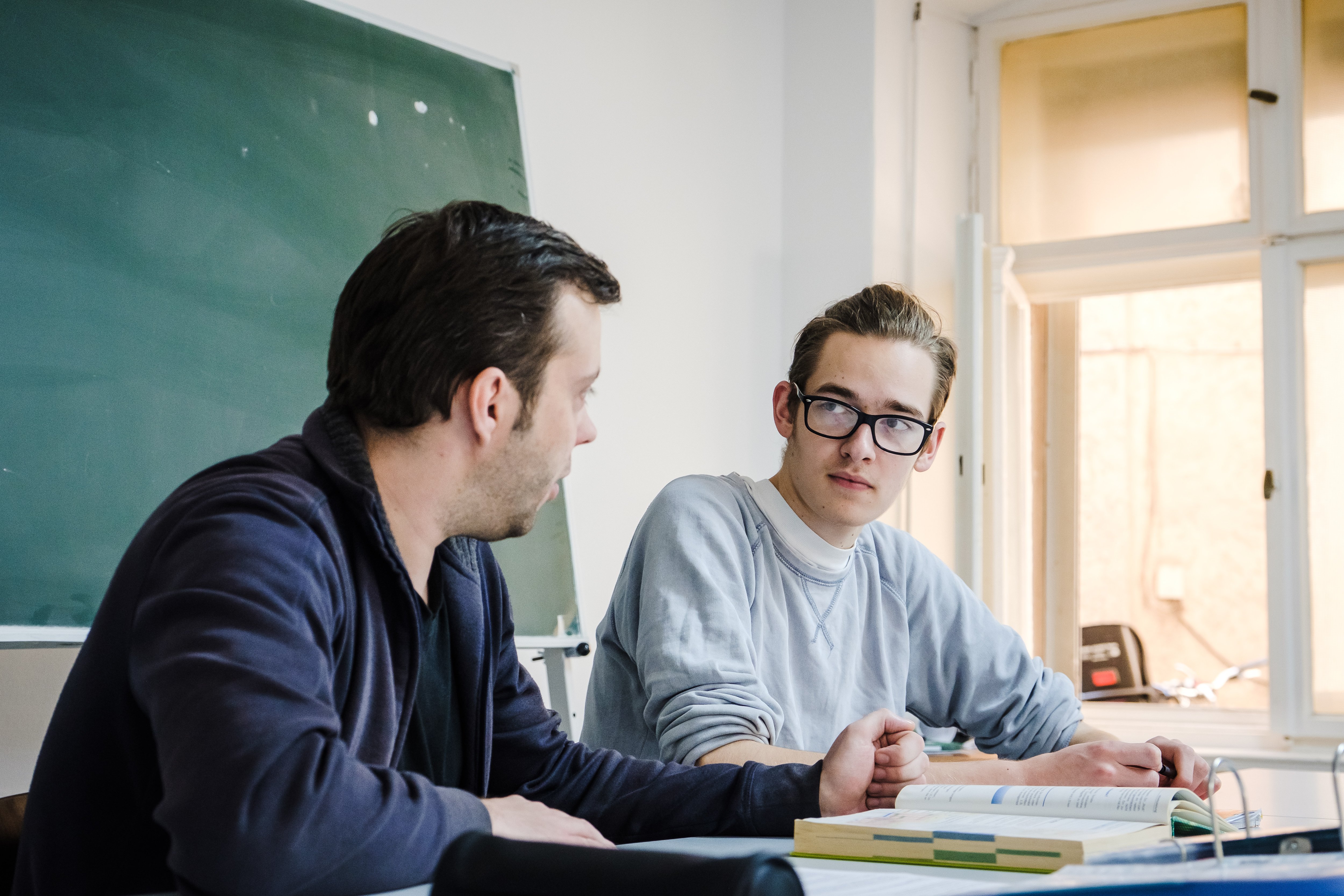 Zwei Männer sitzen an einem Tisch in einem Raum. Der Mann auf der linken Seite hat dunkles Haar und trägt eine dunkle Jacke. Er spricht mit dem Mann auf der rechten Seite, der eine Brille und einen hellgrauen Pullover trägt. Vor dem Mann mit der Brille liegt ein aufgeschlagenes Buch auf dem Tisch. Im Hintergrund ist eine grüne Tafel und ein Fenster zu sehen, durch das Tageslicht in den Raum fällt.