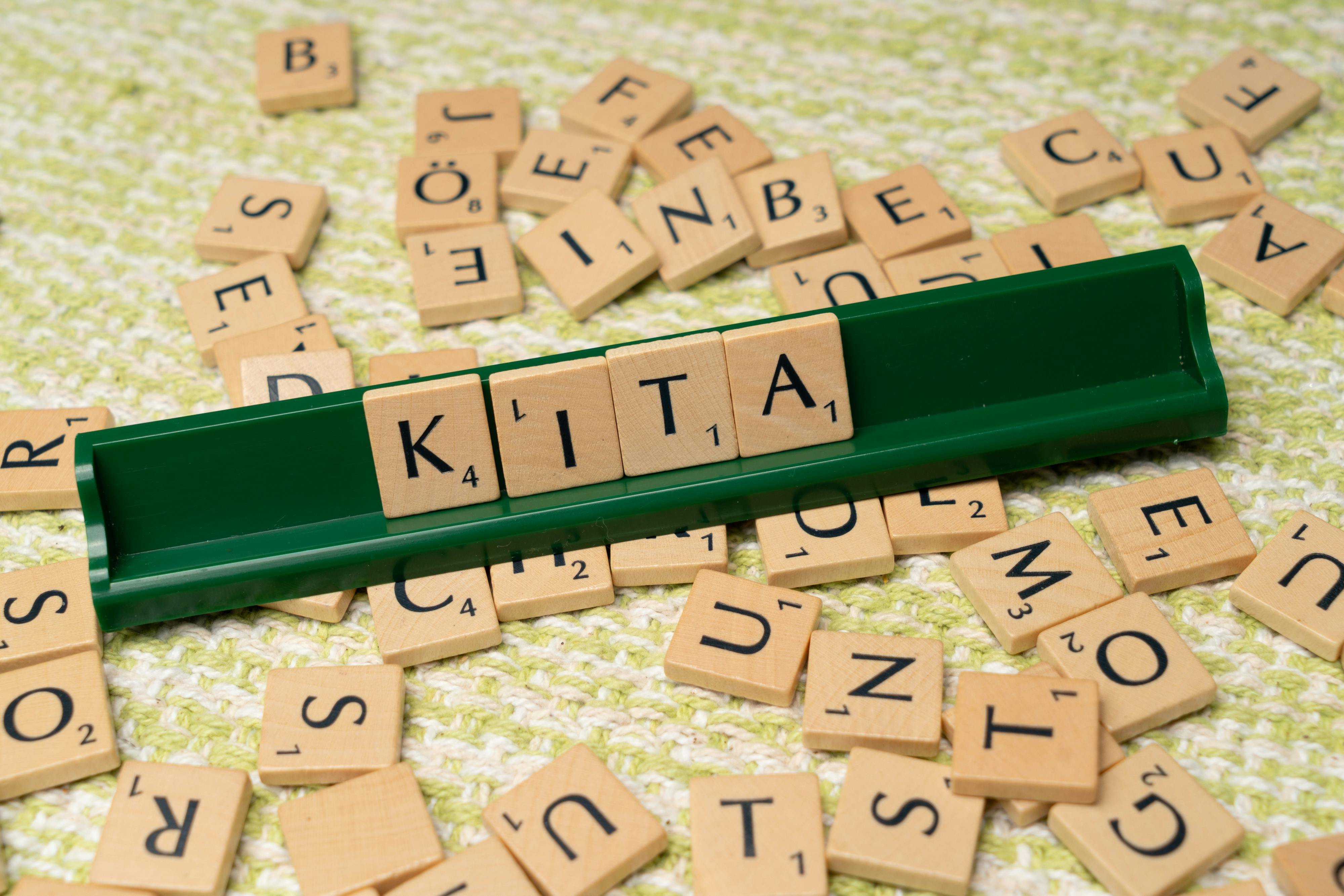 Das Bild zeigt ein Scrabble-Spiel. Auf einem grünen Halter sind die Buchstaben K, I, T und A angeordnet, die das Wort