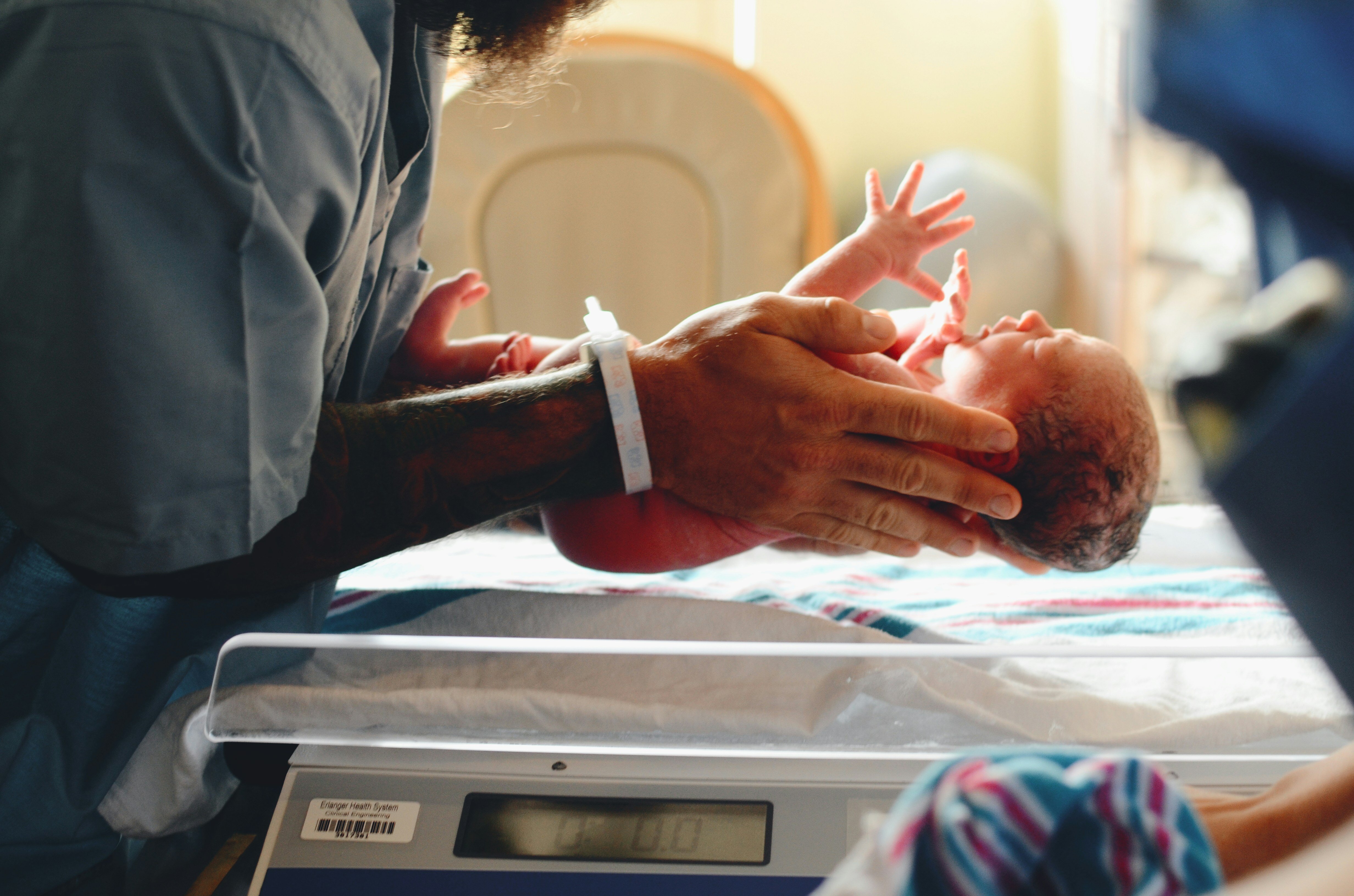 Eine Person hält ein neugeborenes Baby. Die Person trägt ein Krankenhausarmband und hat tätowierte Arme. Das Baby liegt auf einer Decke auf einer Waage, die in einem Krankenhauszimmer zu sein scheint. Das Baby ist nackt und hat noch die typische rötliche Haut eines Neugeborenen. Im Hintergrund ist ein unscharfer Stuhl zu sehen.