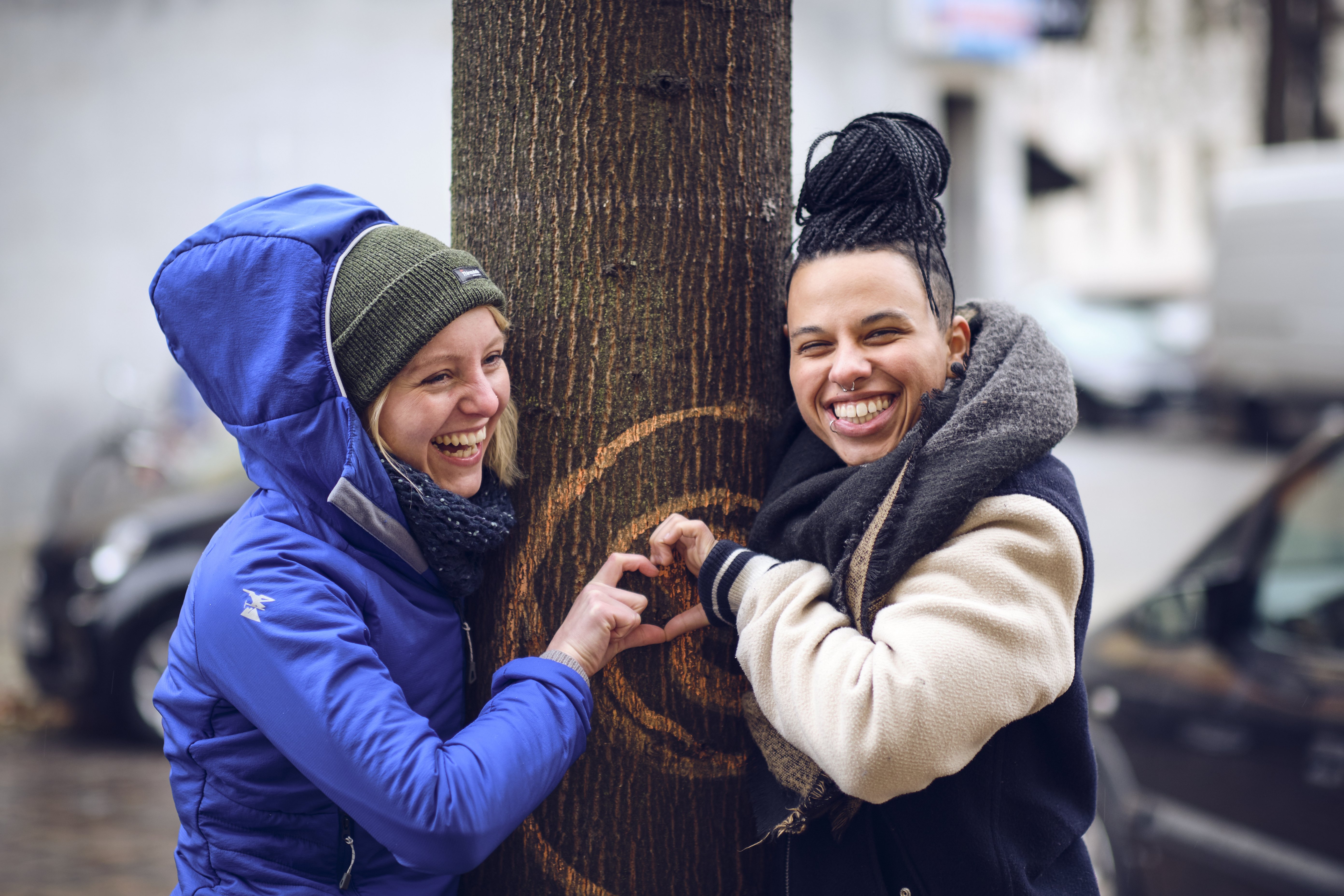 Zwei lachende Personen, die sich um einen Baumstamm herum lehnen. Die Person links trägt eine blaue Jacke mit Kapuze und eine grüne Mütze. Die Person rechts hat geflochtene Haare und trägt eine helle Jacke mit einem dunklen Schal. Beide Personen formen mit ihren Händen ein Herz vor dem Baumstamm. Der Baum hat Kreise, die mit Kreide oder Farbe darauf gezeichnet sind. Im Hintergrund sind verschwommen Autos und Gebäude zu sehen.
