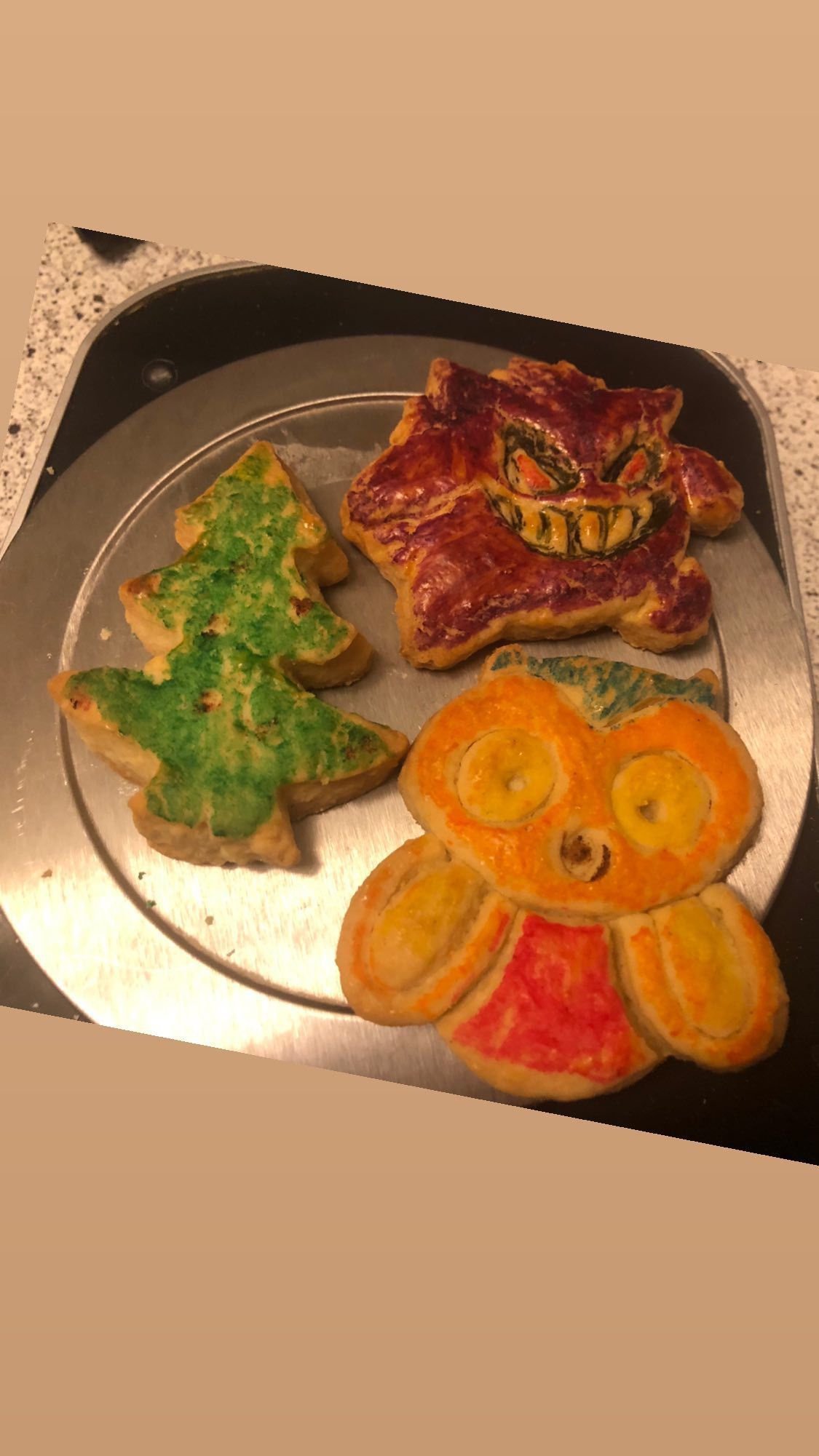 Drei bunte Kekse auf einem runden Metalltablett. Der Keks oben rechts hat die Form eines Gangers und ist lila mit einem gruseligen Gesicht darauf. Der Keks oben links ist in Form eines Weihnachtsbaums und grün gefärbt. Unten rechts befindet sich ein Keks in Form einer Eule, die orange und gelb bemalt ist.