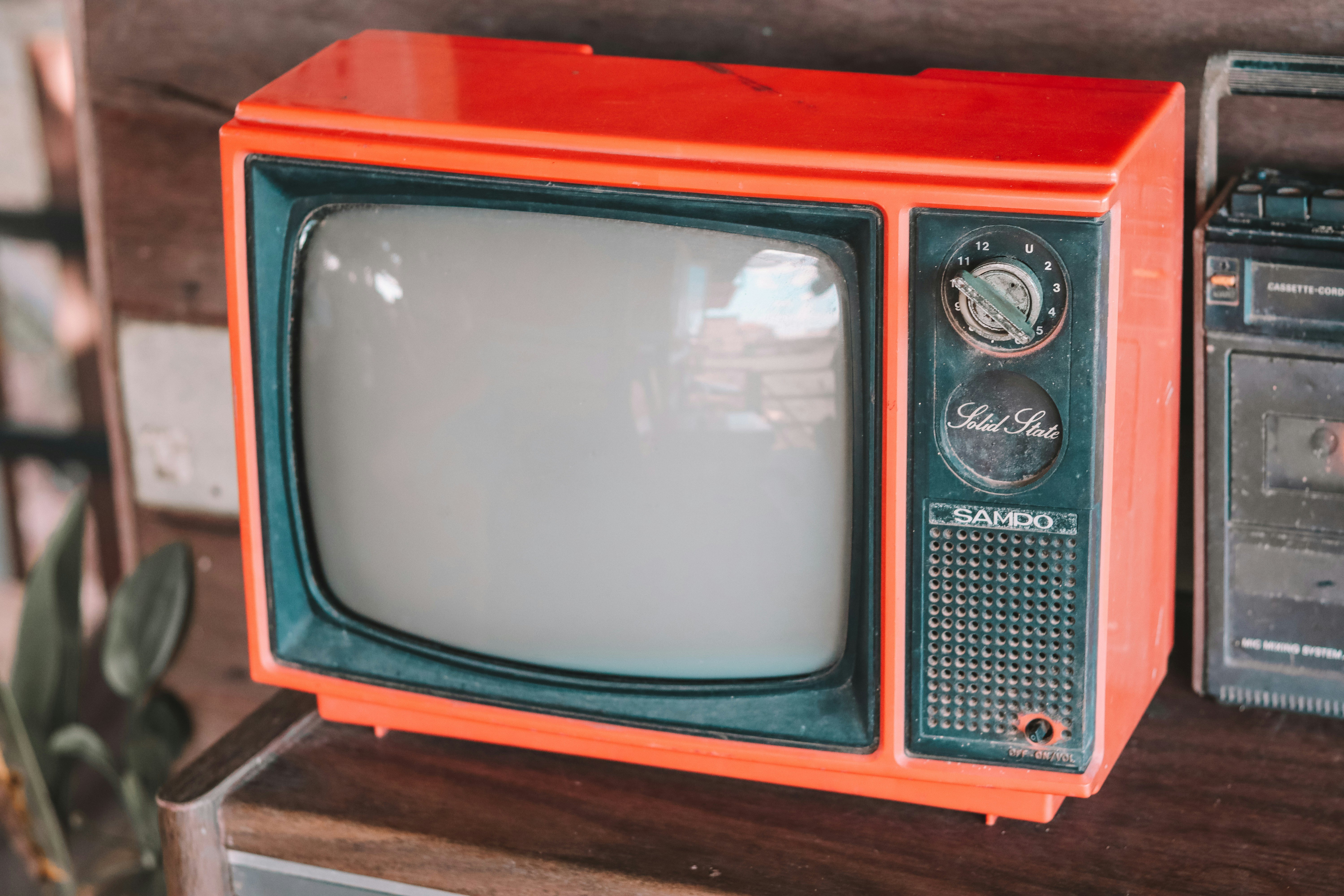 Ein alter Fernseher mit einem orangefarbenen Gehäuse. Der Bildschirm ist leicht gewölbt und hat einen breiten schwarzen Rahmen. Rechts neben dem Bildschirm befinden sich zwei Drehknöpfe und ein Lautsprechergitter. Auf dem Fernseher steht