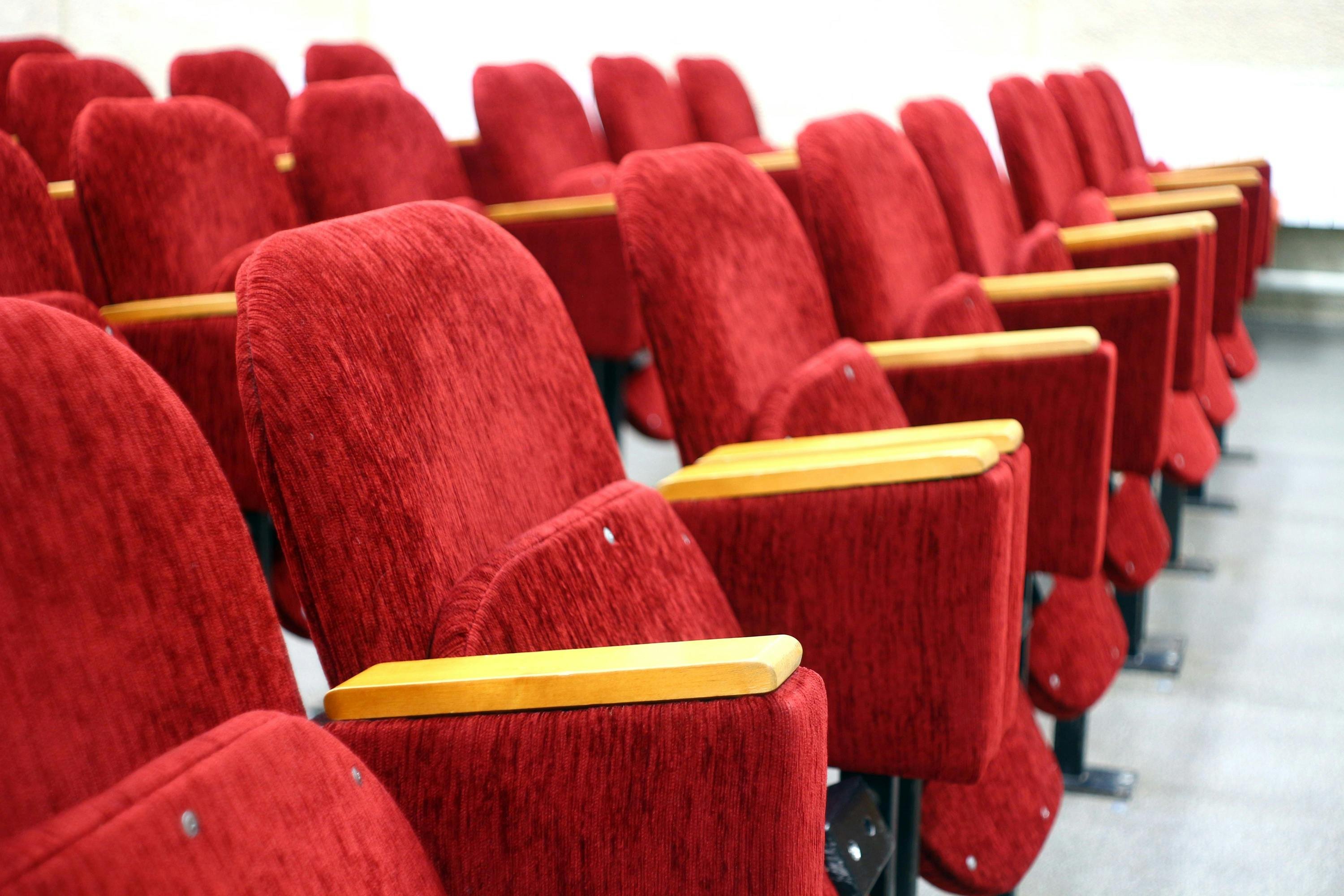 Eine Reihe von roten Theatersitzen. Die Sitze sind mit rotem Stoff bezogen und haben hölzerne Armlehnen. Sie sind in einer Linie angeordnet und scheinen in einem Auditorium oder einem Theater zu stehen. Der Boden ist grau und die Sitze sind auf Metallgestellen montiert. Die Perspektive des Bildes ist seitlich, sodass die Sitze in einer diagonalen Linie verlaufen.