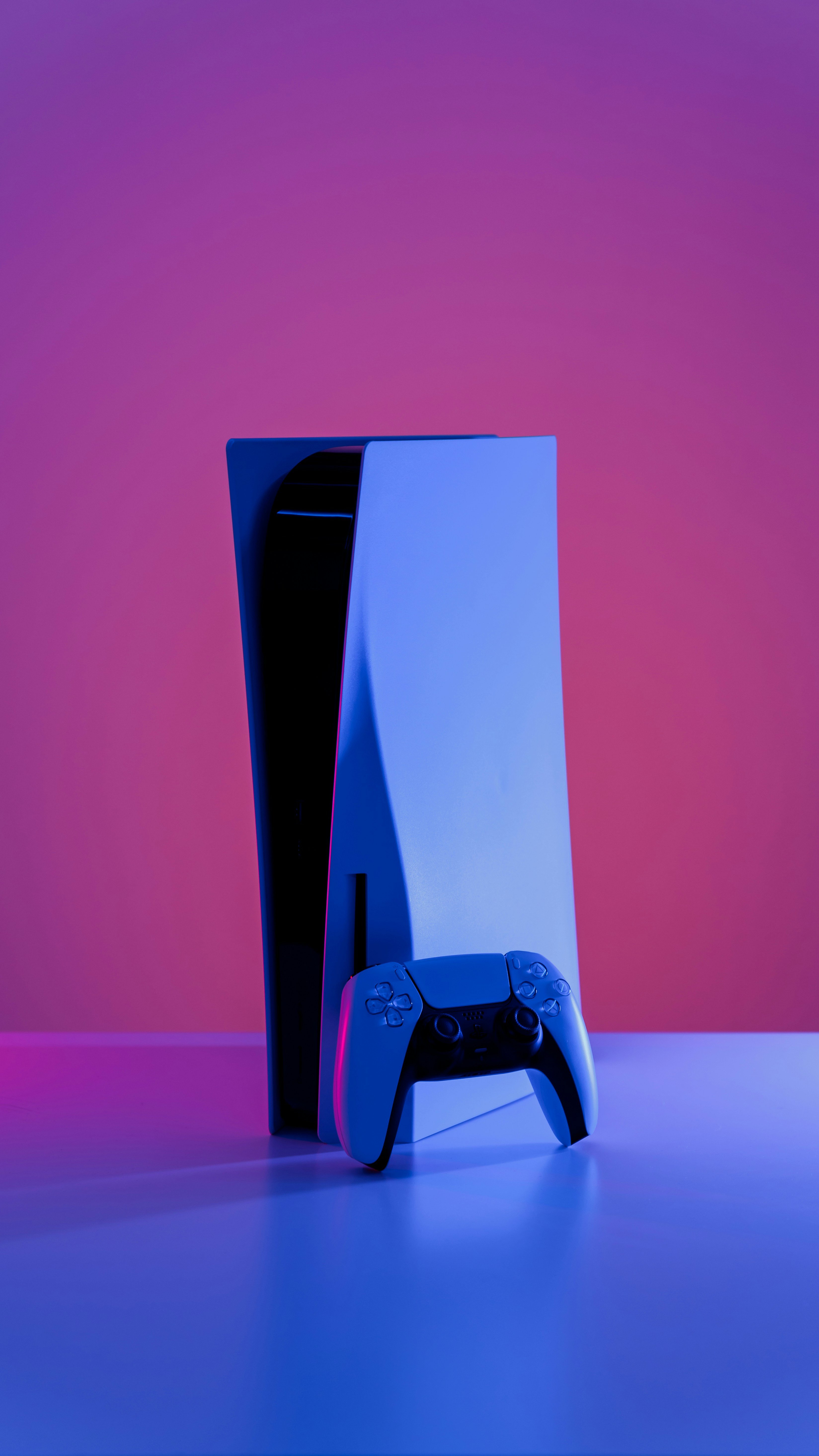 Eine PlayStation 5 Konsole mit einem DualSense-Controller davor. Die Konsole steht aufrecht auf einer Oberfläche. Der Hintergrund ist in einem Farbverlauf von Rosa zu Lila gehalten, und die Konsole sowie der Controller sind in blauem Licht beleuchtet, was einen futuristischen Effekt erzeugt.