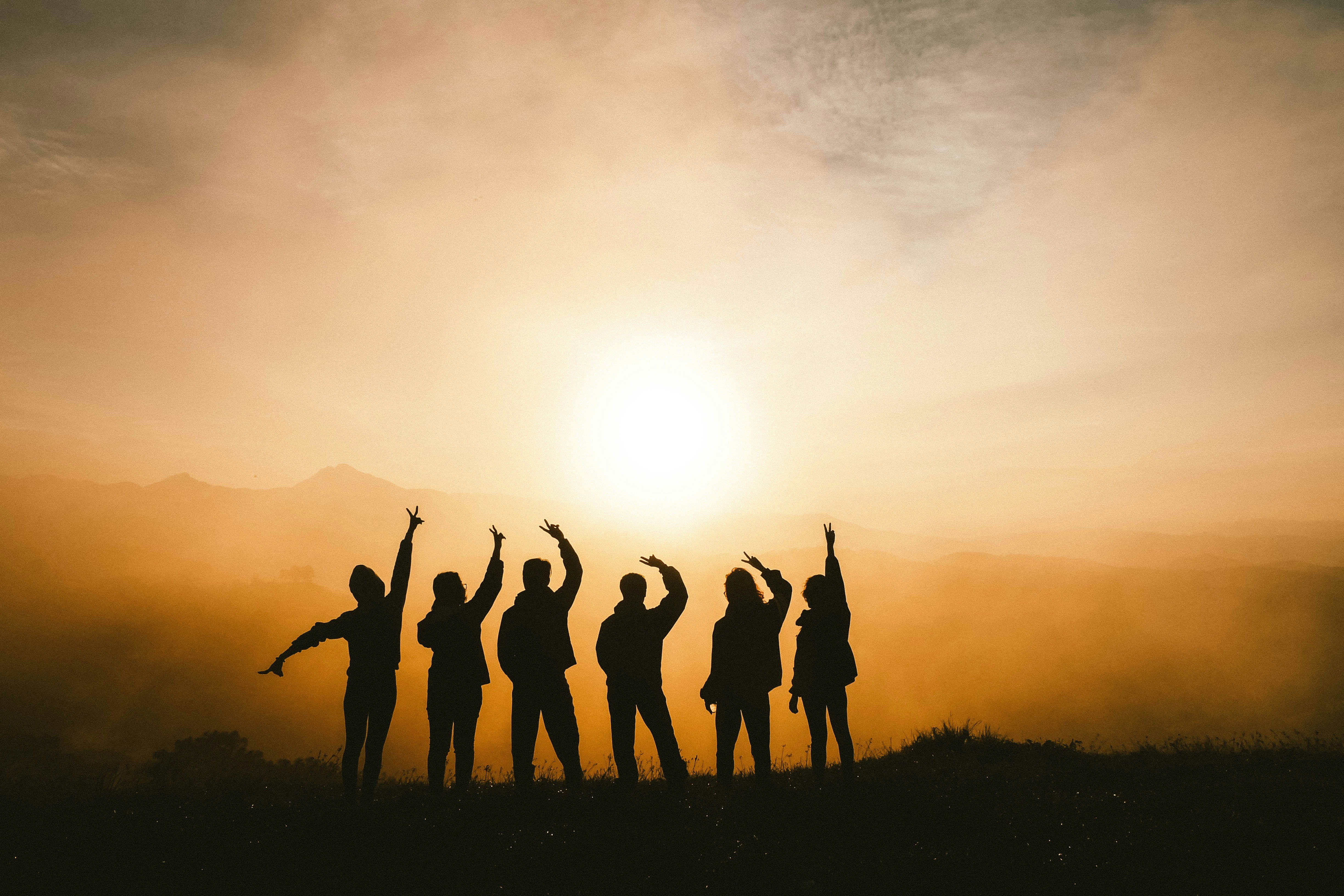 Silhouetten von sieben Personen, die auf einem Hügel stehen. Sie sind vor einem Sonnenuntergang positioniert, wobei die Sonne tief am Himmel steht und einen warmen, goldenen Schein über die Szene wirft. Einige von den Personen strecken die Arme in die Luft. Der Hintergrund ist neblig. Die Landschaft im Hintergrund ist hügelig.